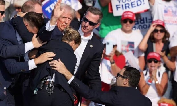 Donald Trump waving hand with a bloodied face while he was assisted by Secret Service personnel after he was shot in the right ear during a campaign rally